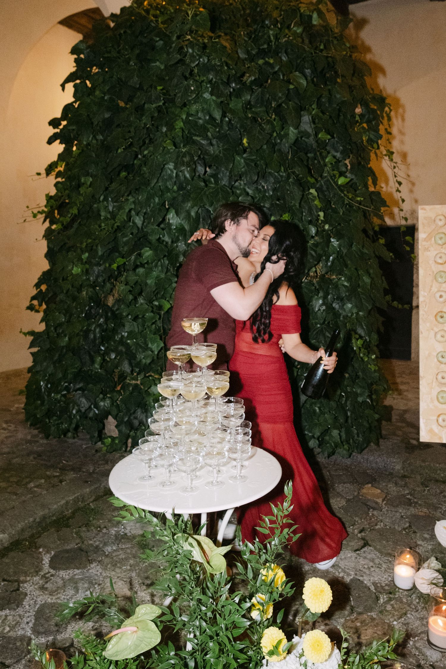 Champagne tower setup with flowers and greenery against stone wall at evening reception.