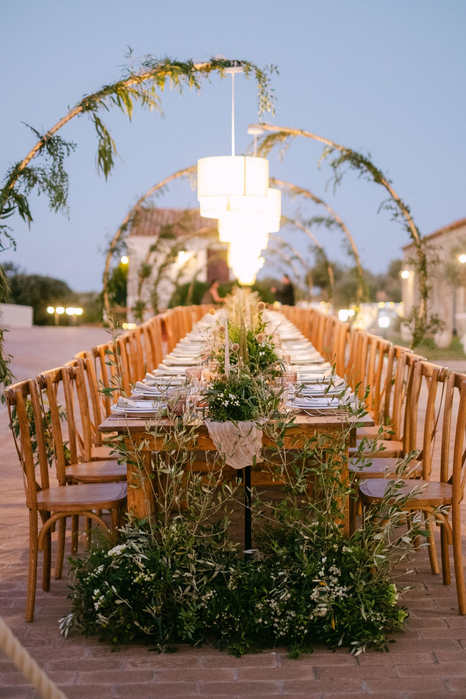 An outdoor wedding reception setup with wooden chairs and floral arrangements under hanging lights.