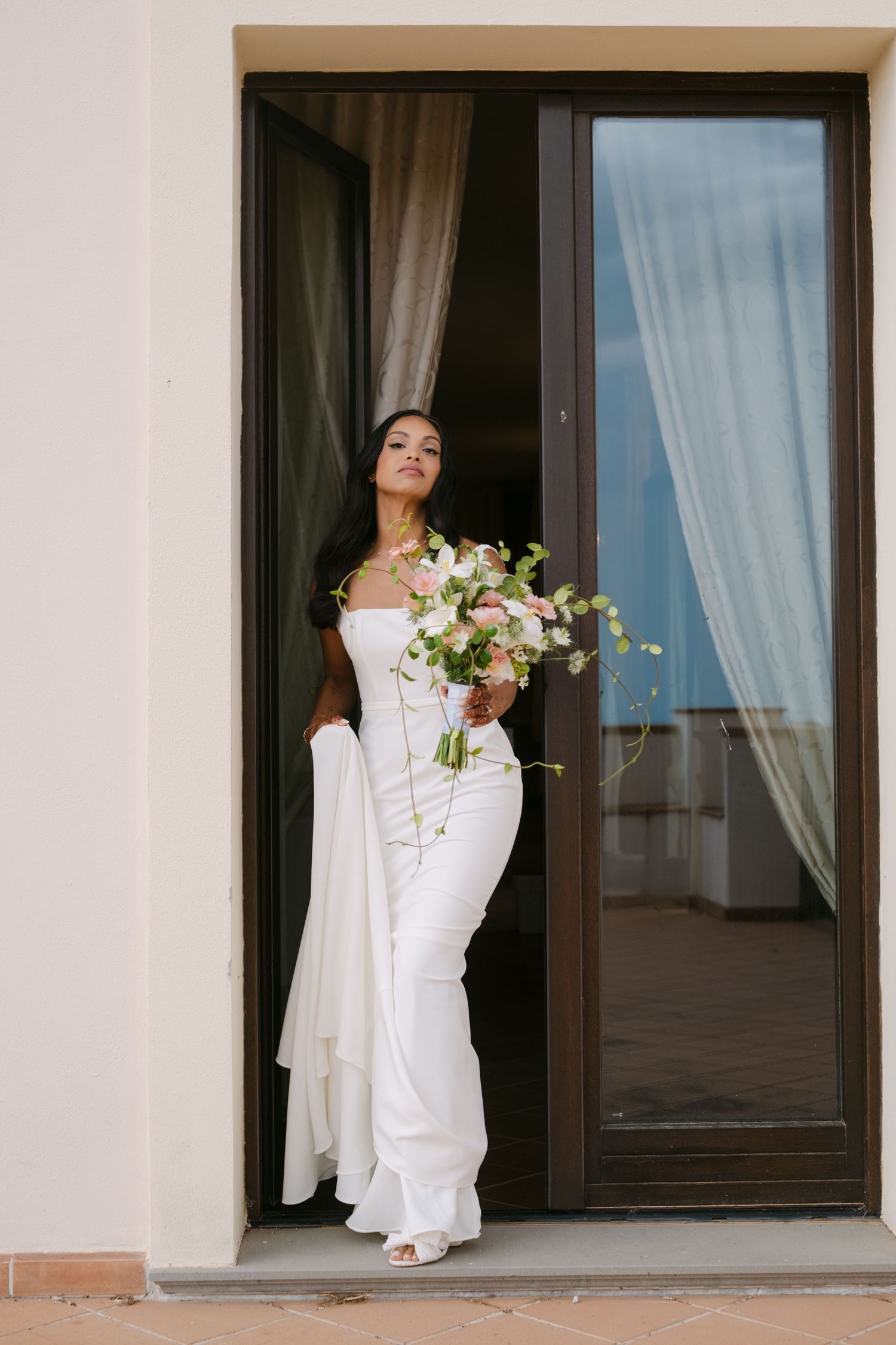 Bride in white dress poses elegantly by window with floral bouquet.