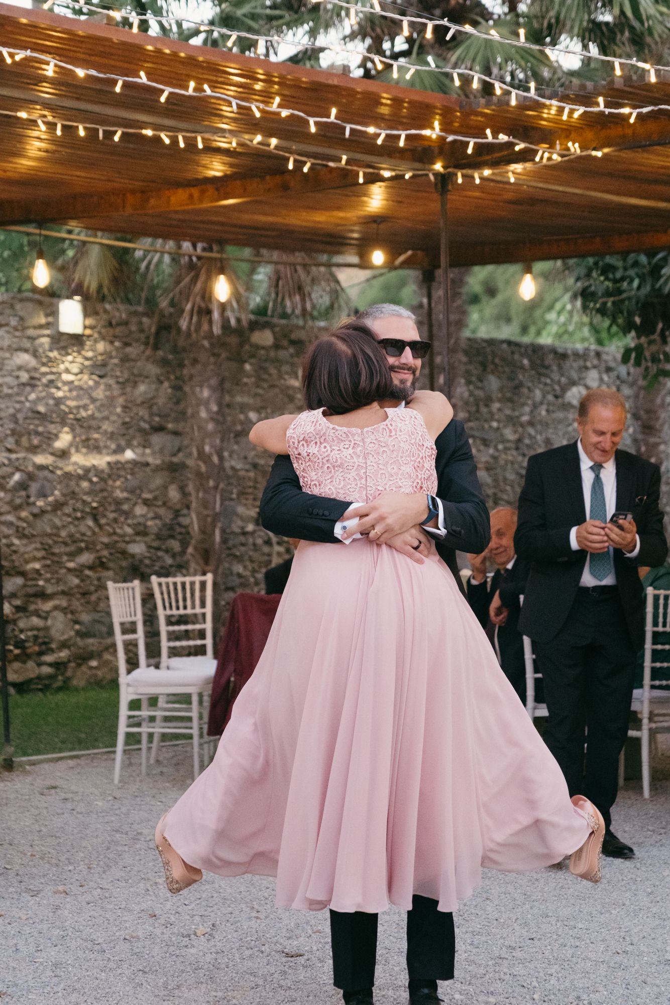People in formal attire dance and embrace at an outdoor venue decorated with string lights and stone walls.