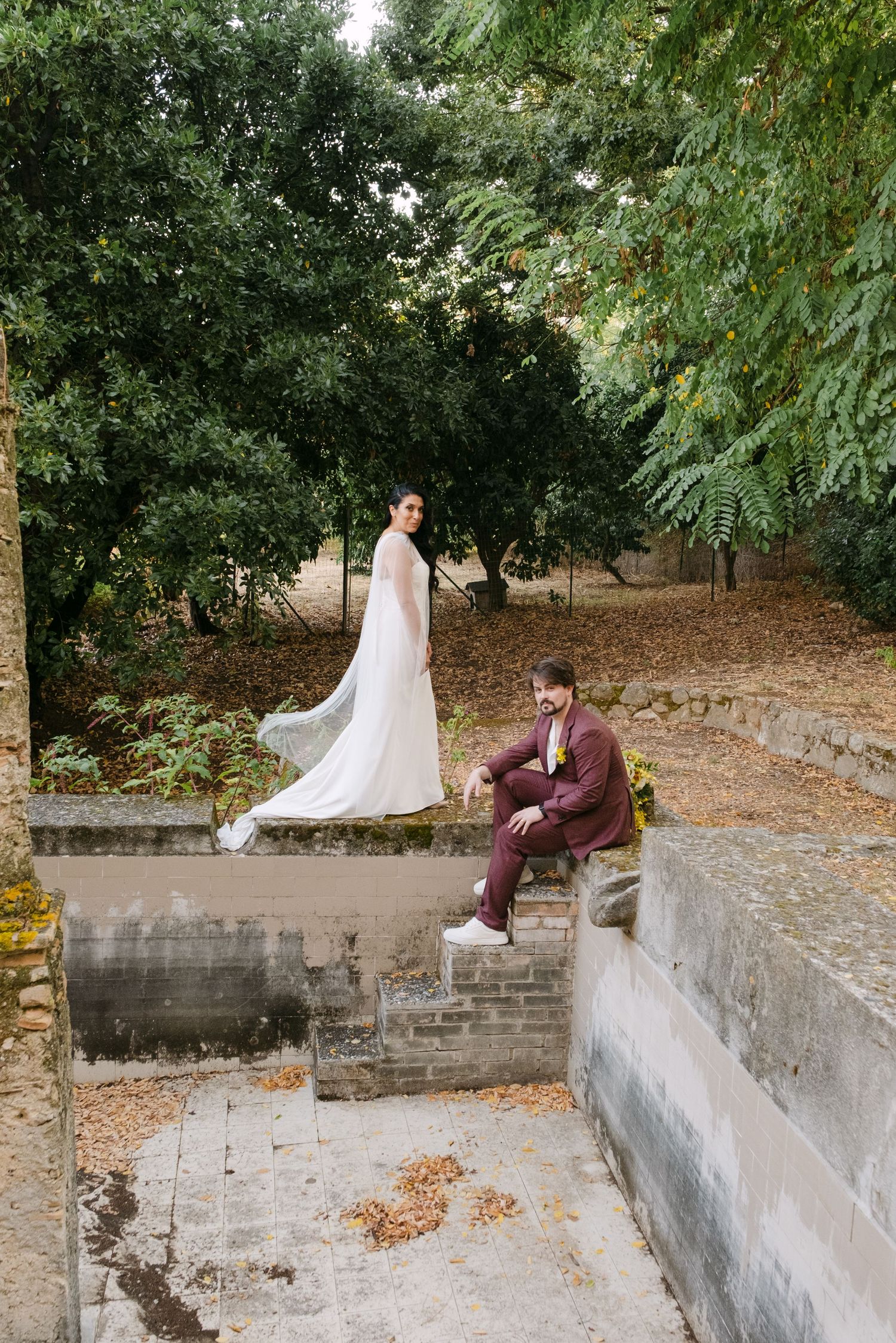 Wedding couple poses playfully on stone steps surrounded by lush greenery in an outdoor garden setting.