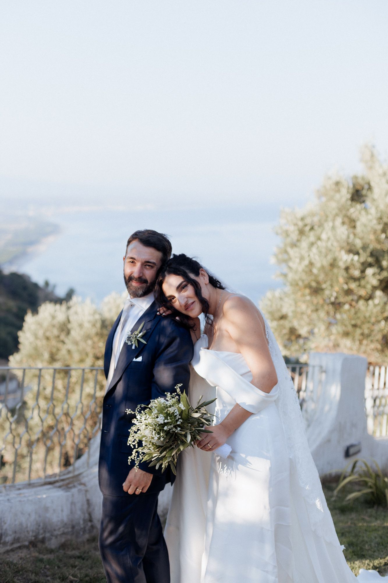 Bride and groom embrace on hillside terrace overlooking coastal view.