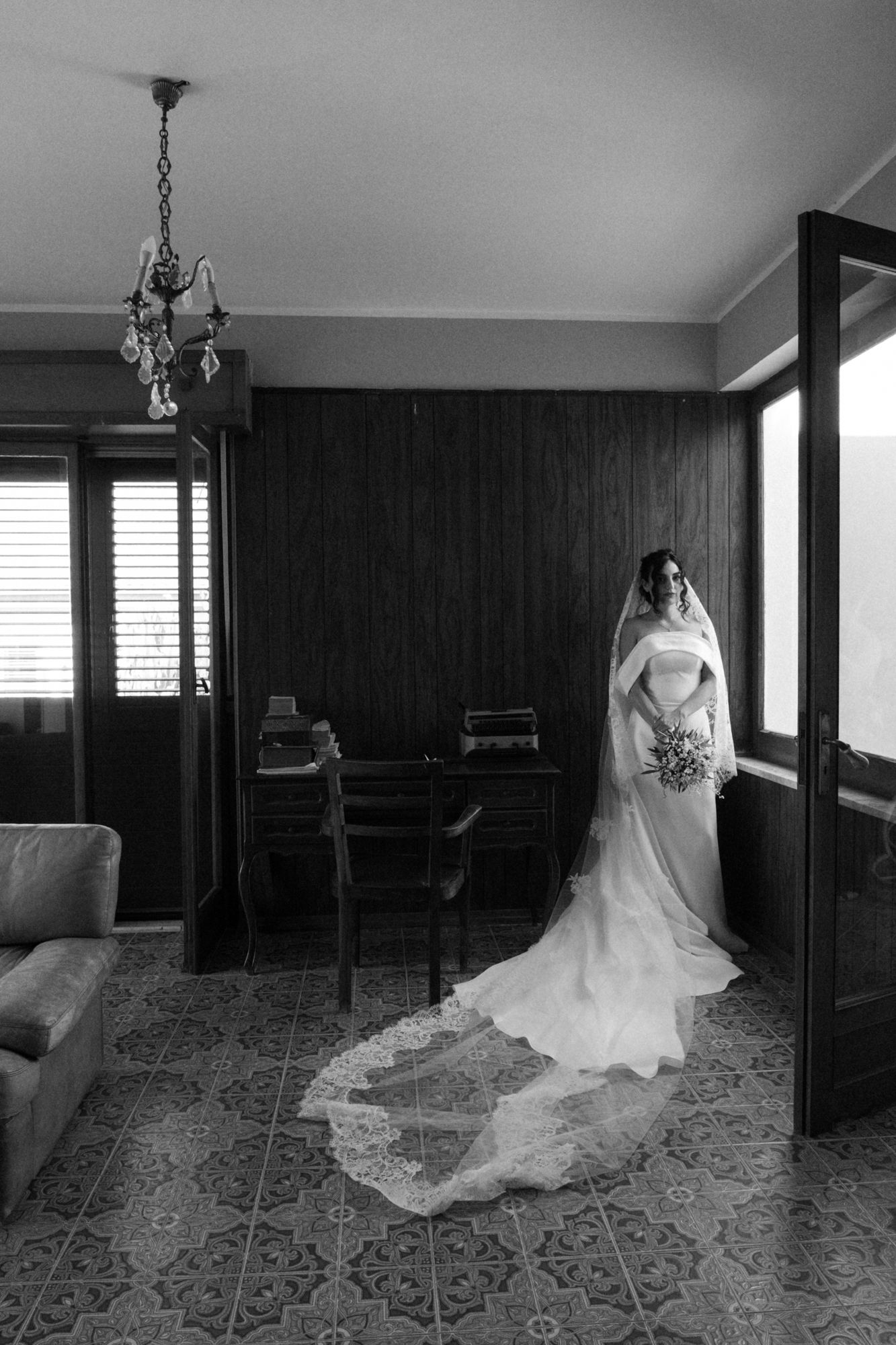 A dramatic black and white photo of a bride in a flowing wedding gown and veil standing by a window.