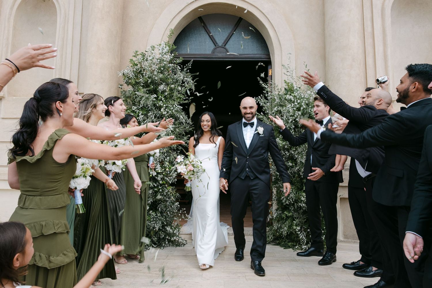 Wedding party celebrates with outstretched arms as newlyweds exit church ceremony between greenery-adorned archway.