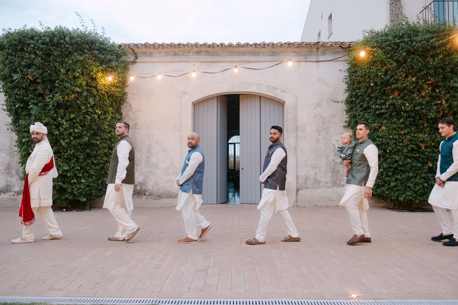 A line of people in light colored attire walk in formation outside a vine-covered building at dusk.