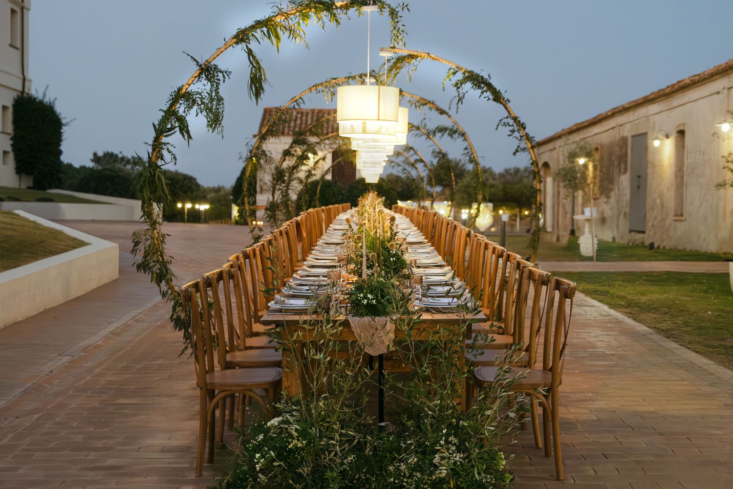 Elegant outdoor wedding reception table set with wooden chairs and hanging floral arrangements at dusk.