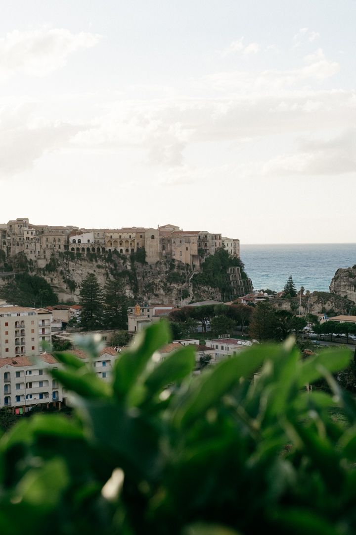Coastal Italian town perched on dramatic cliffs overlooking the Mediterranean Sea with green foliage in foreground.