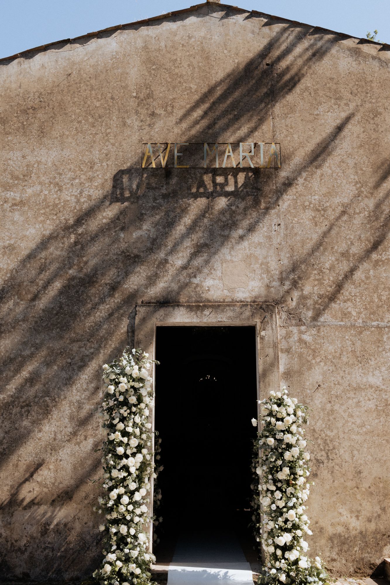 White floral arrangements frame a dark doorway against a textured beige wall with dramatic shadows.