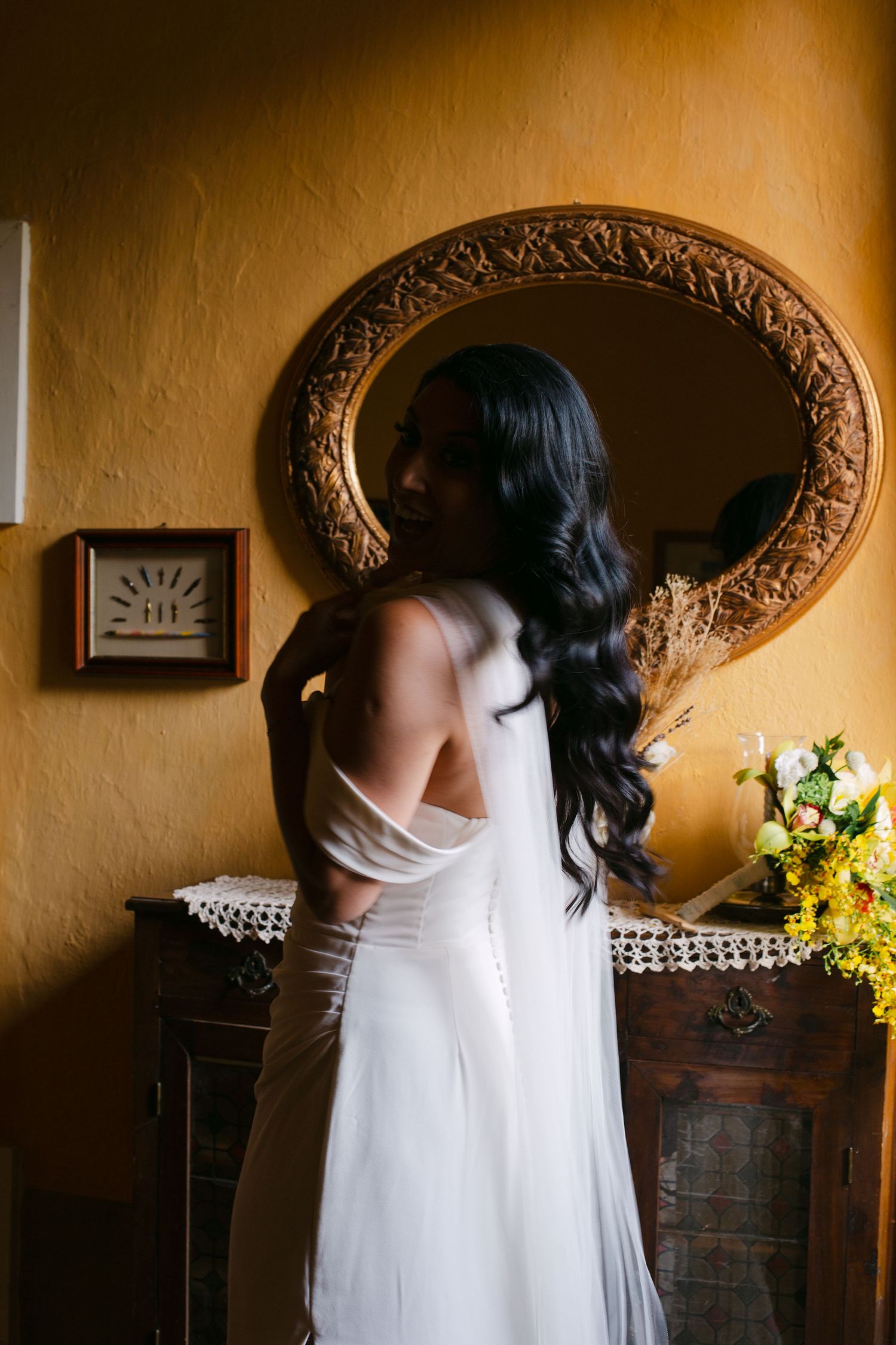A flowing white wedding dress captured from behind against warm yellow walls and ornate mirror.