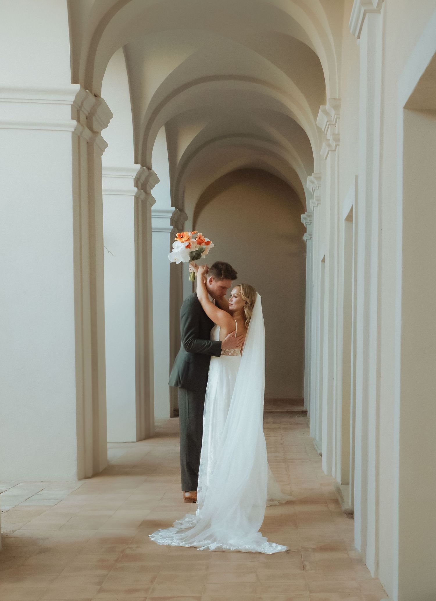 Romantic wedding couple embraces in an elegant white arched hallway with classical architecture.
