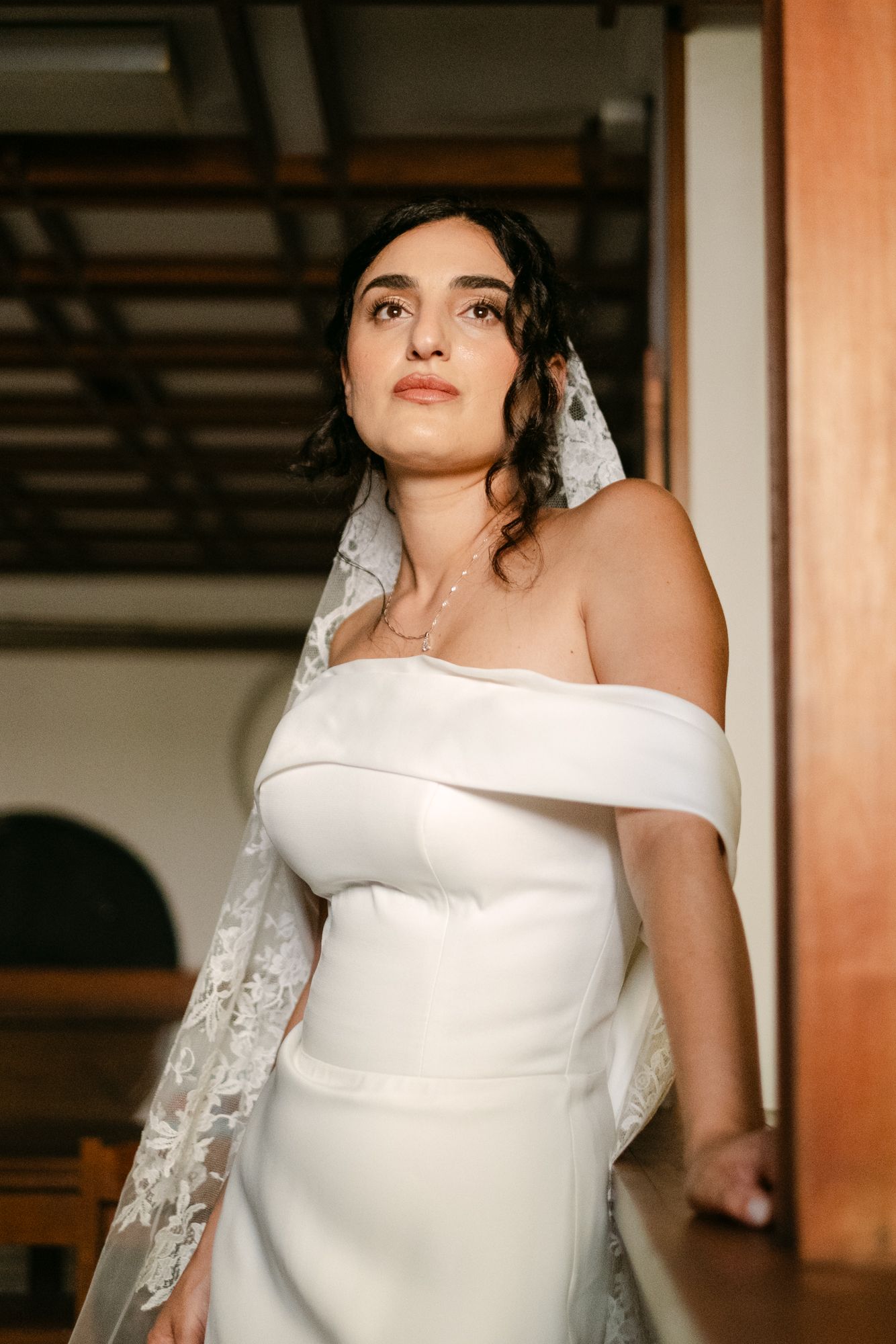 Elegant off-shoulder white wedding dress with lace veil details in warm-toned indoor setting.