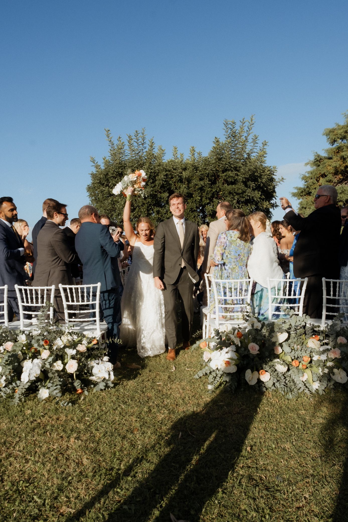Wedding guests gather outdoors on a grassy hill during sunset as long shadows stretch across the ground.