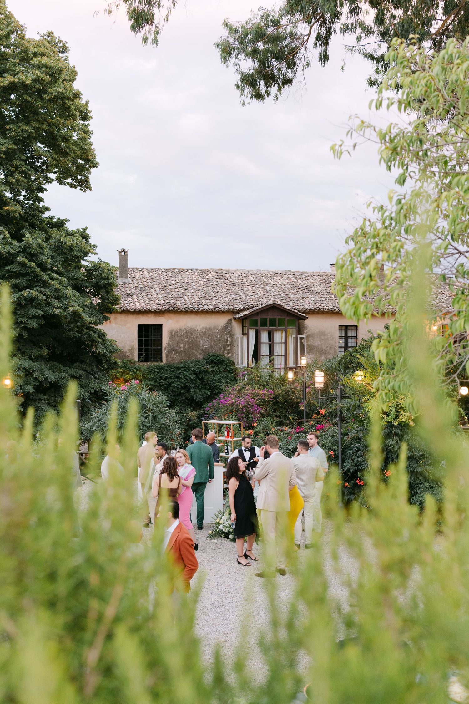 Wedding guests gather on a garden path leading to a rustic villa.
