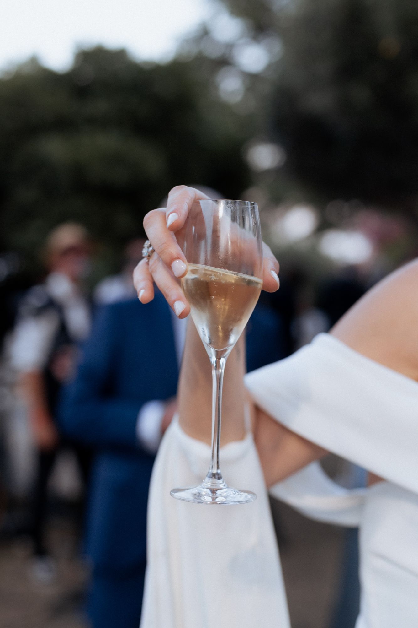 A delicate hand holding a champagne flute against a blurred outdoor background.