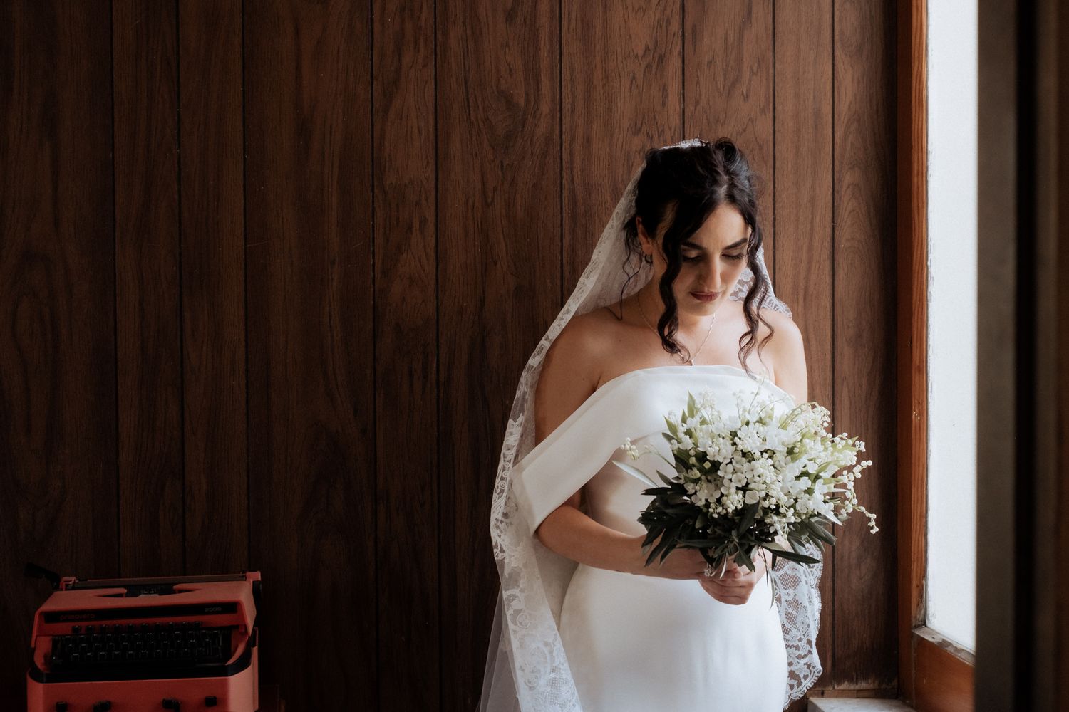 A bride in white holds a bouquet while posing against dark wood paneling.