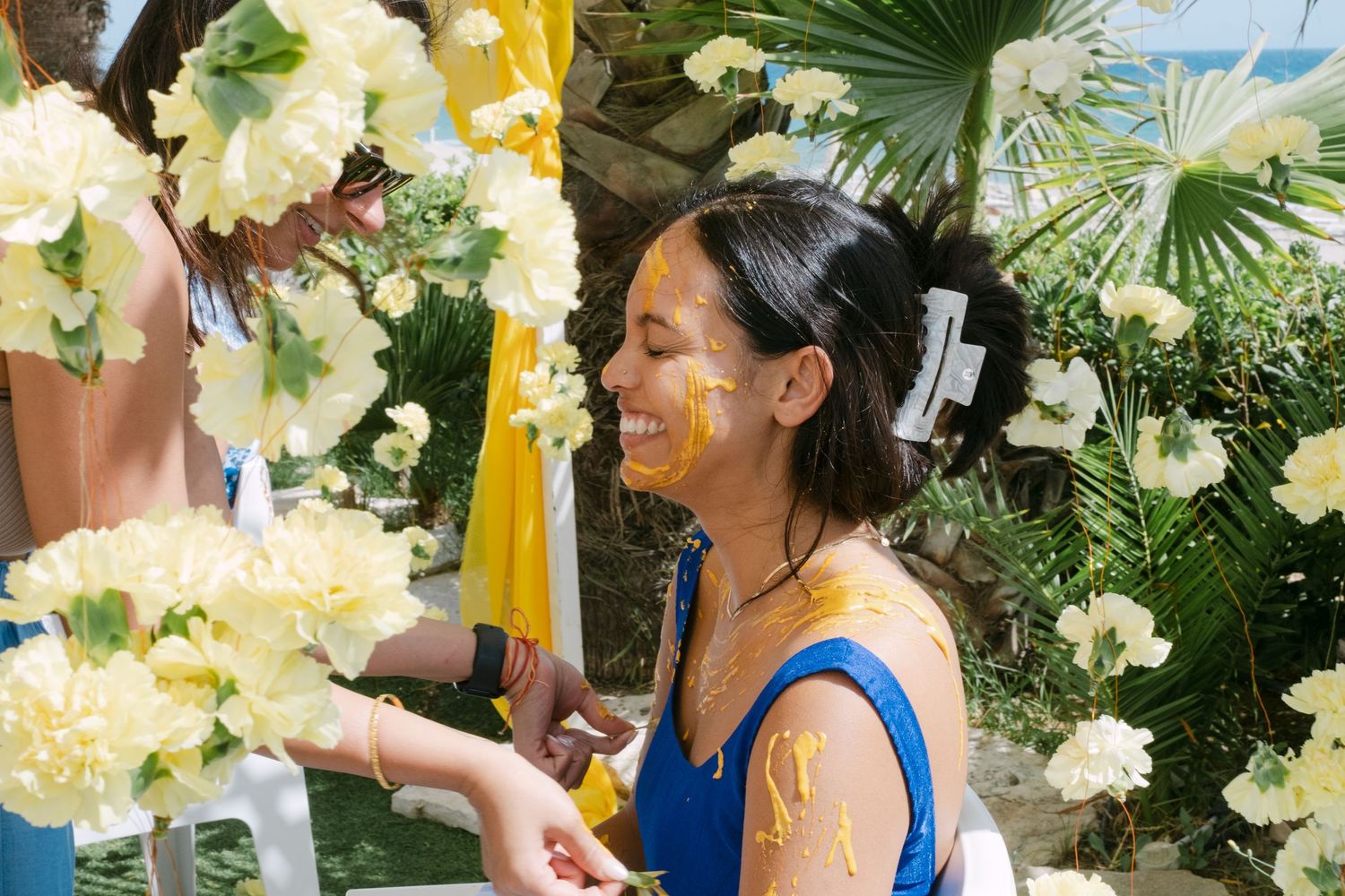 A person in a blue dress smiles while arranging pale yellow flowers among tropical foliage and decor.