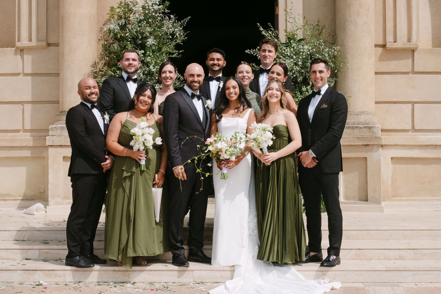 Wedding party in olive green dresses and black tuxedos poses together outside elegant building.