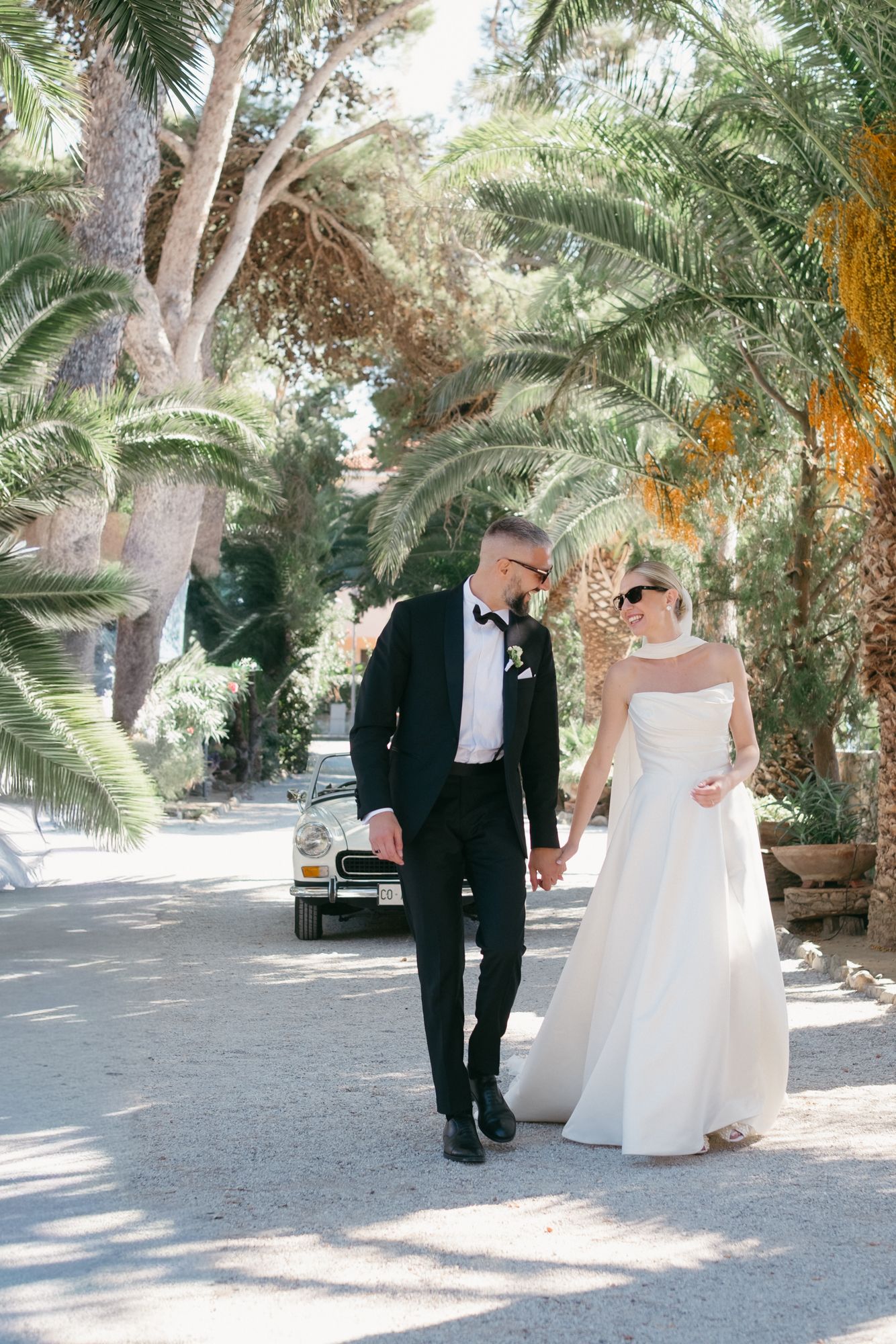 Couple walking together on a palm tree-lined path during a sunny outdoor wedding celebration.