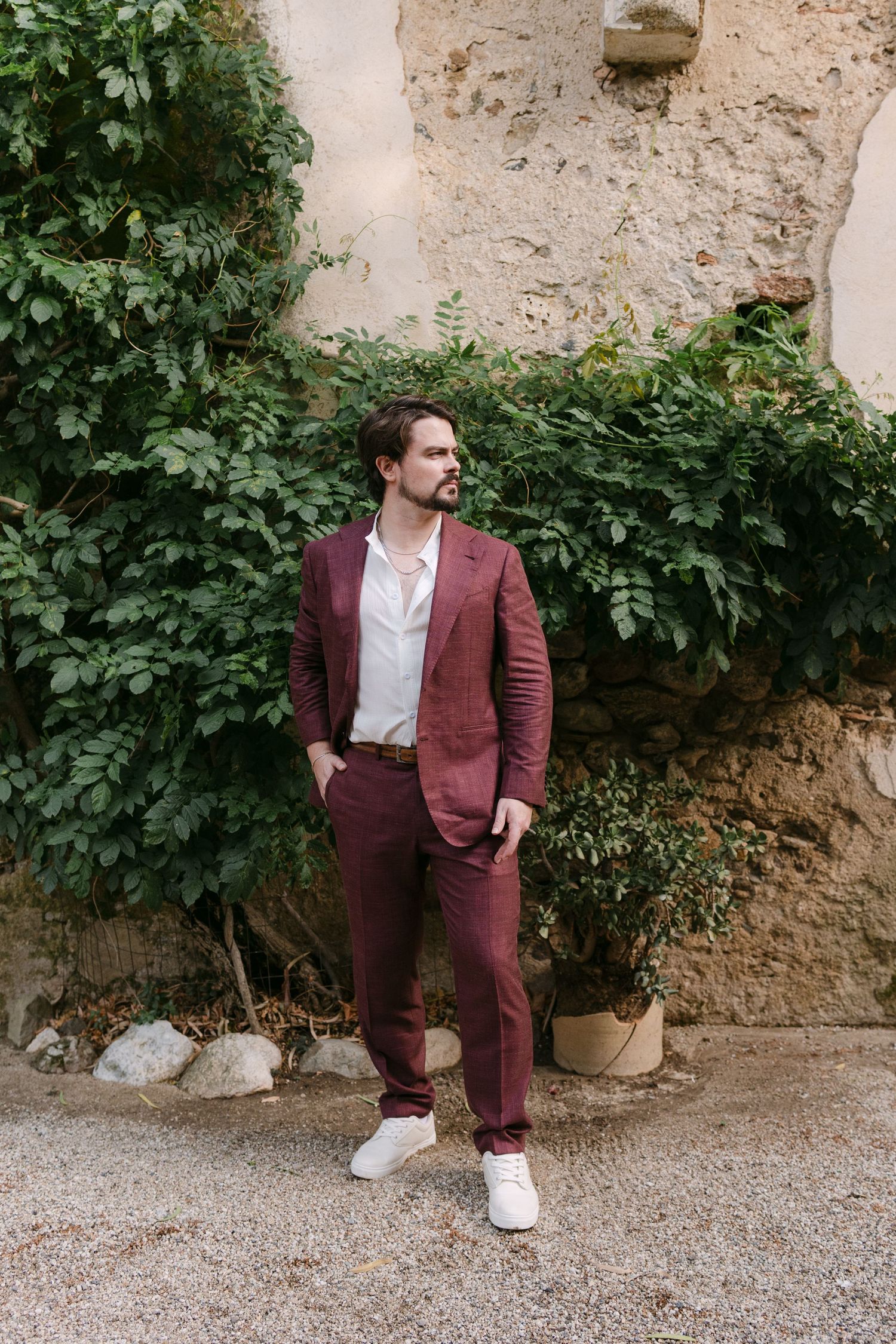 Person in burgundy suit poses against textured stone wall with green foliage.