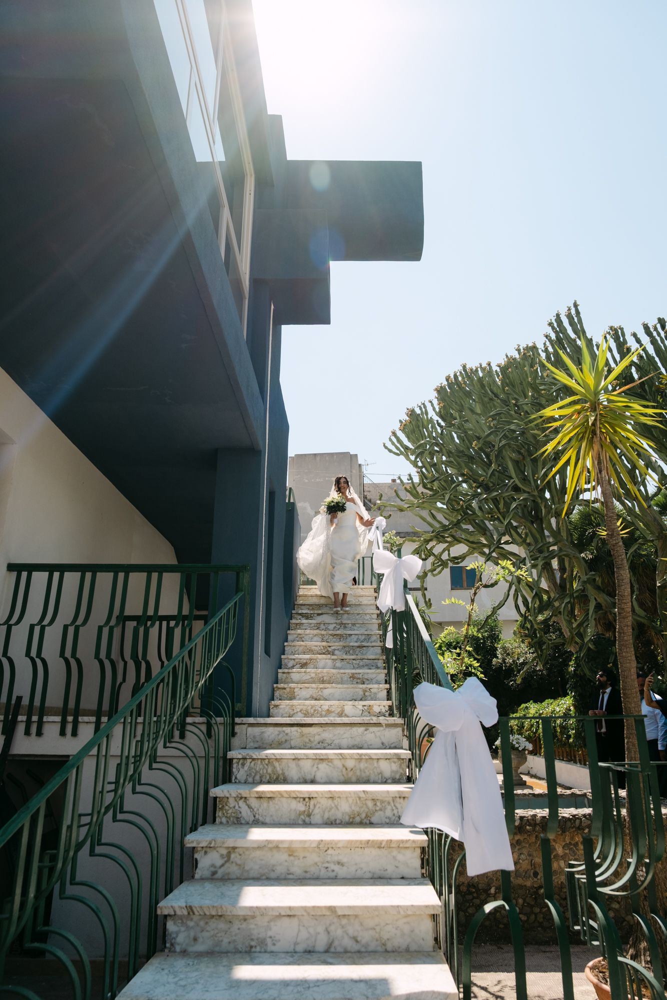 People in white attire ascending outdoor marble stairs with green metal railings and tropical plants.