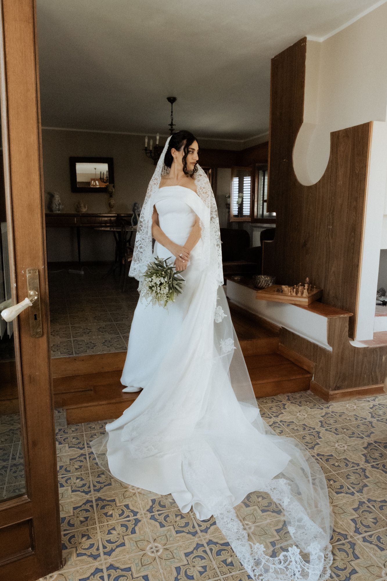 Full-length shot of bride in off-shoulder wedding gown with flowing train on patterned tile floor.