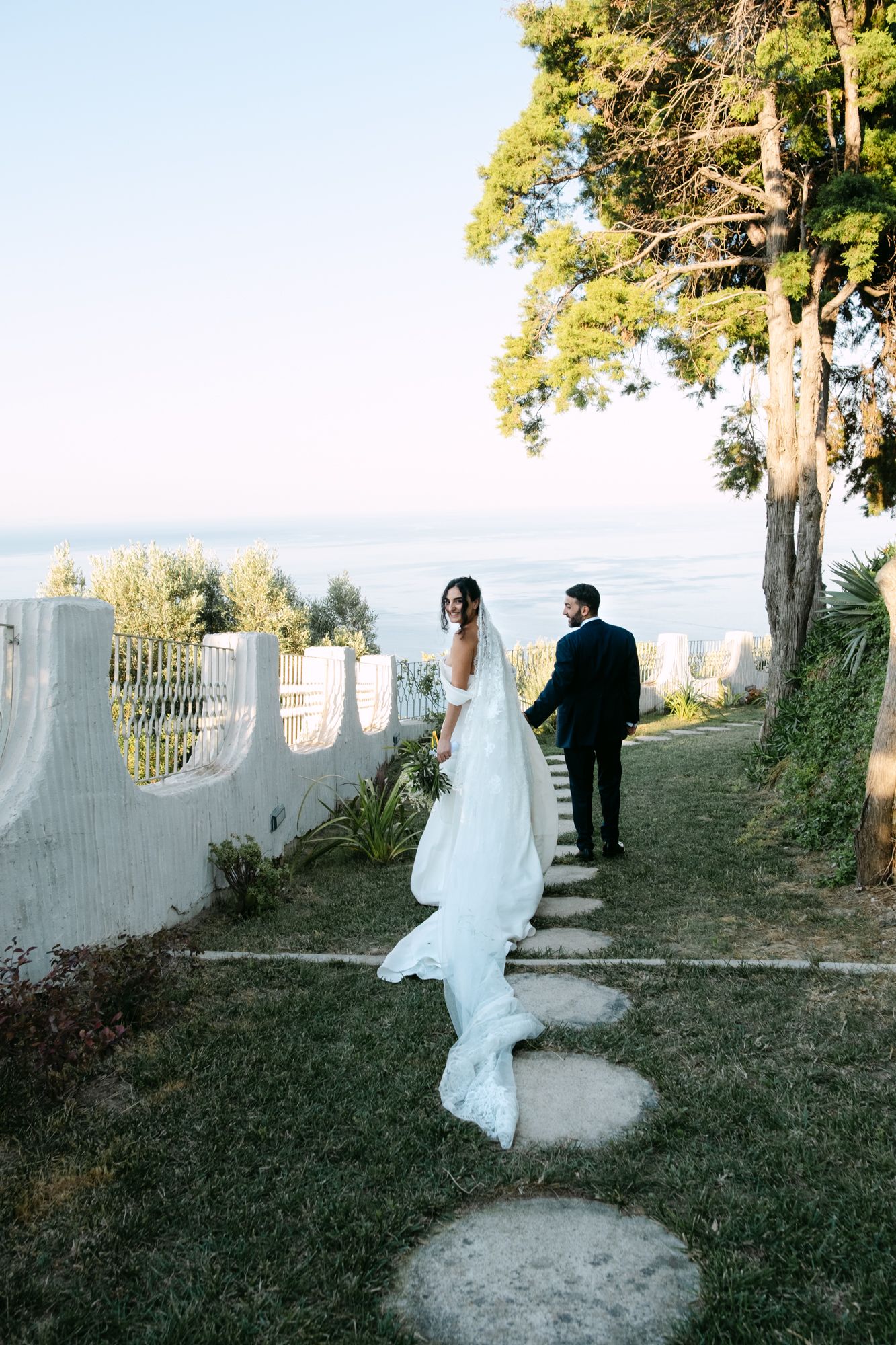 Newlyweds walking down garden path with ocean view and white wall in background.