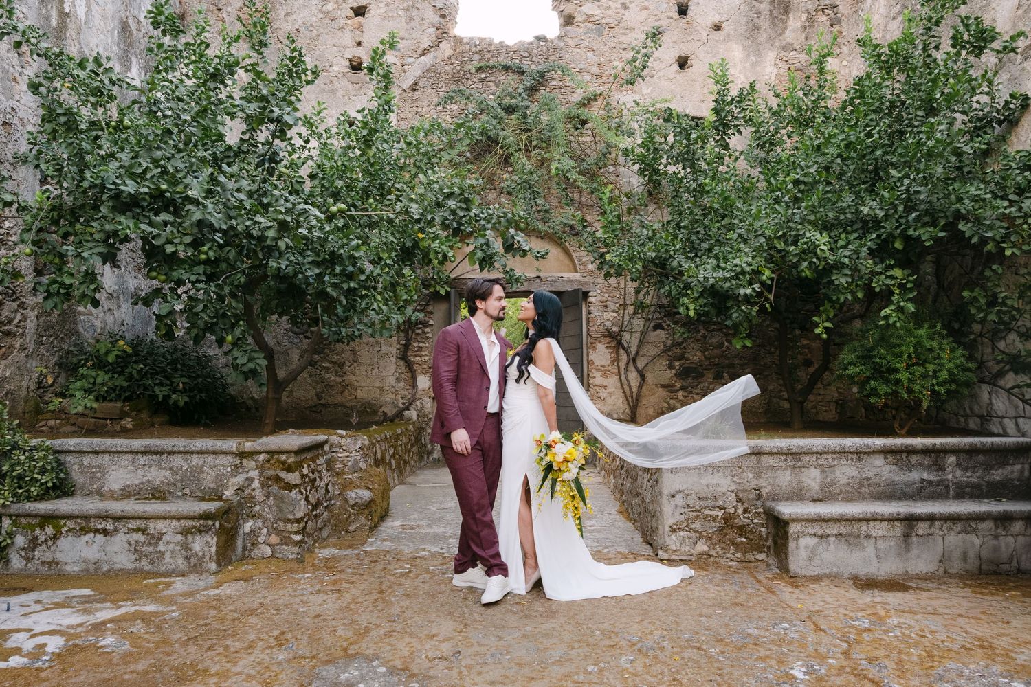 A couple poses for wedding photos in a garden with stone walls and trees.