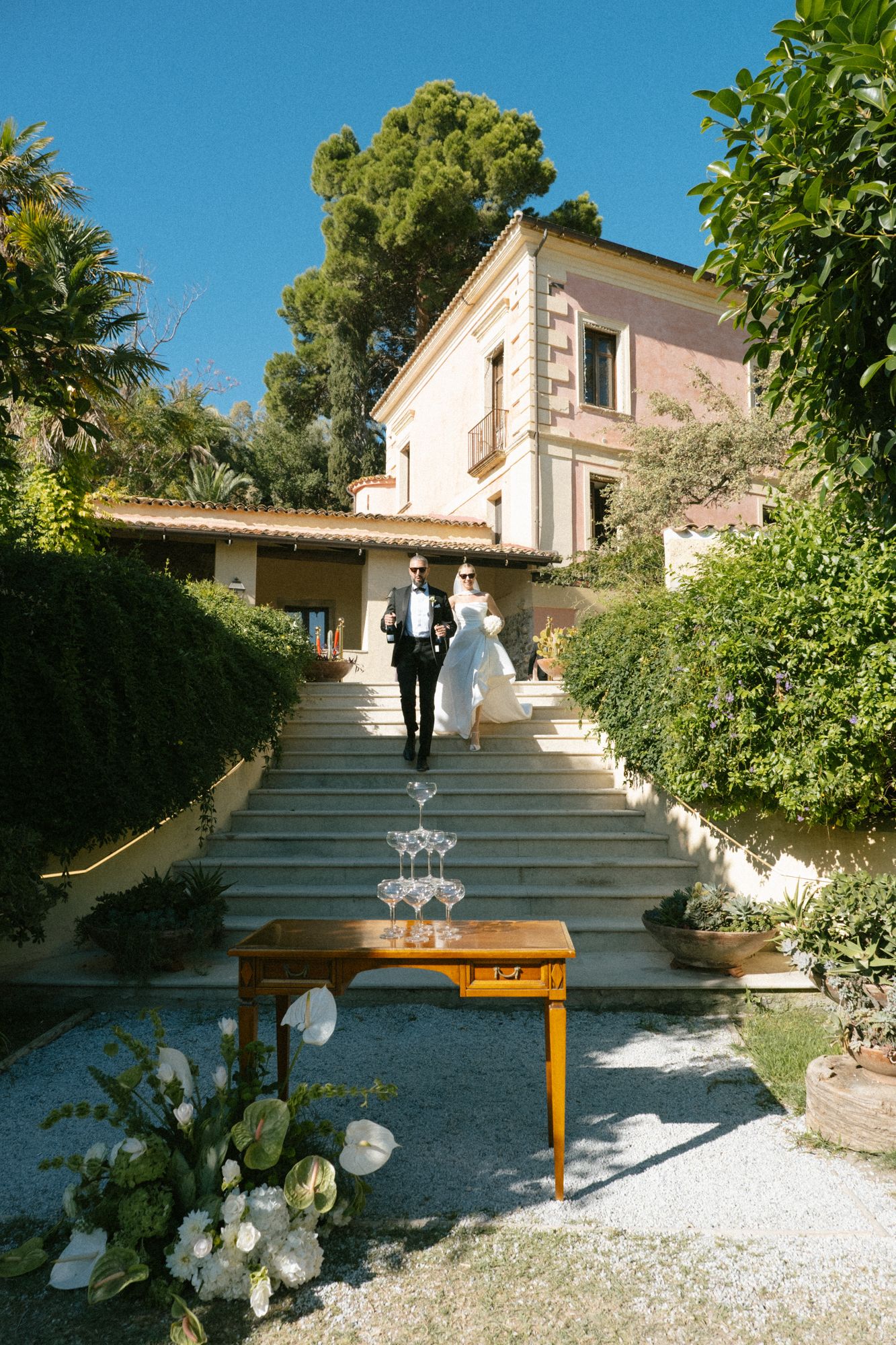 White stone steps decorated with floral arrangements lead up to a Mediterranean-style villa on a sunny day.