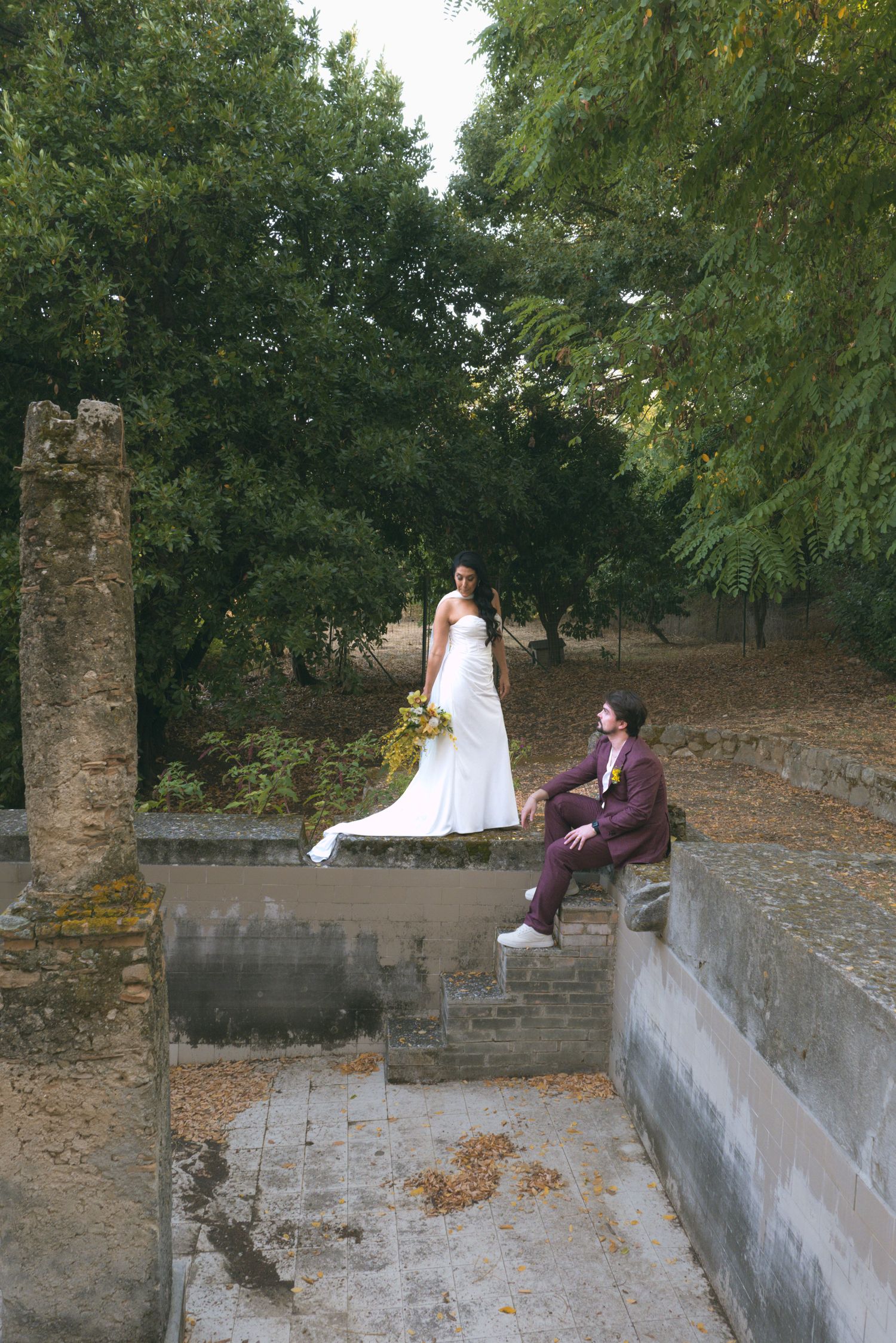 A wedding couple sits on an old stone wall surrounded by lush greenery in an outdoor setting.