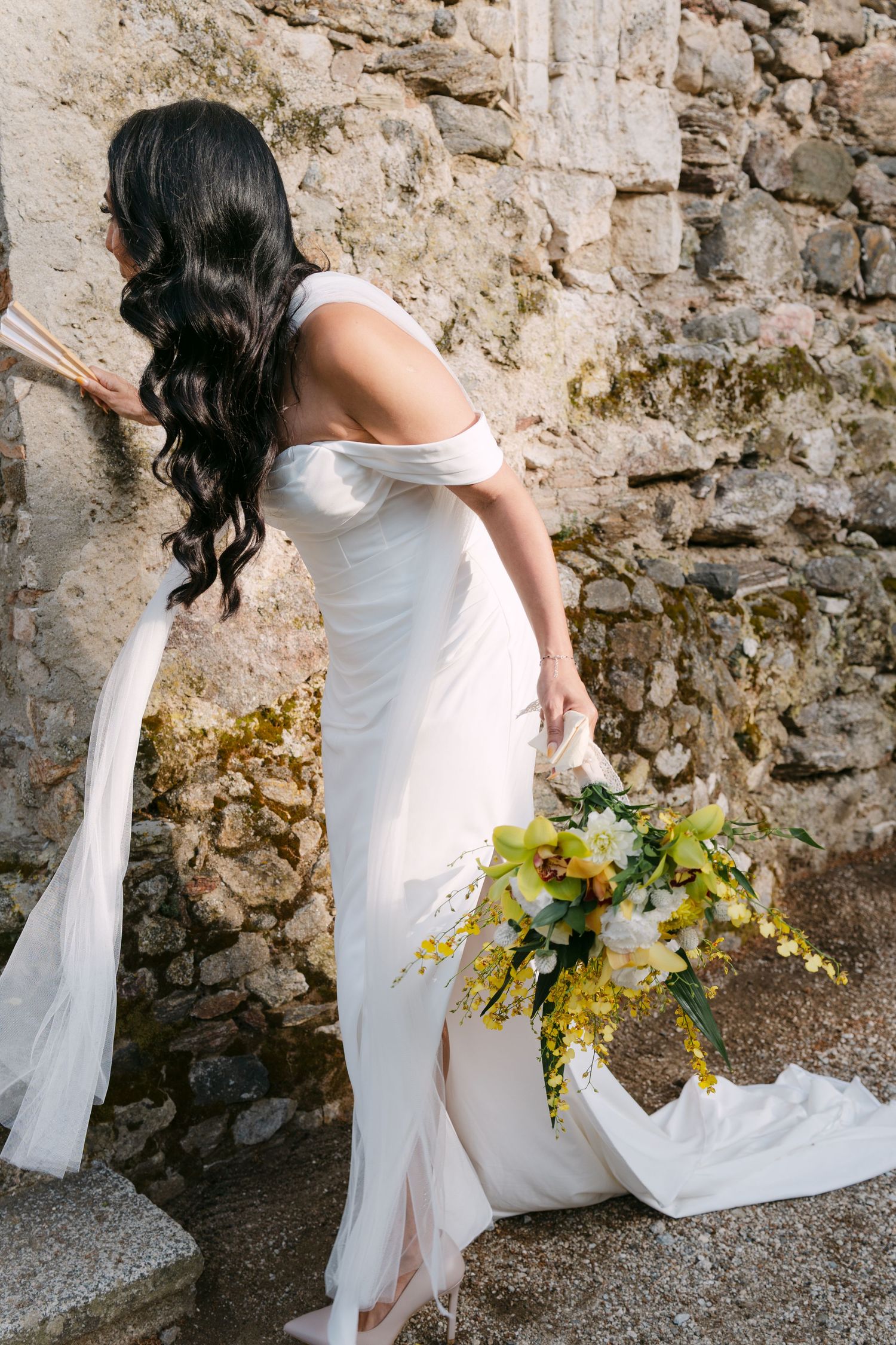 A bride in a white dress holds a yellow bouquet against a rustic stone wall backdrop.