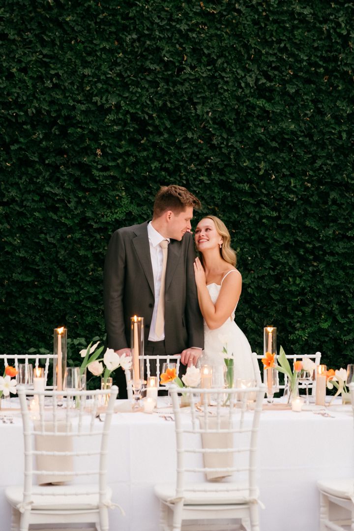 An elegant outdoor wedding reception setup with white chairs and tables against a green hedge wall backdrop.