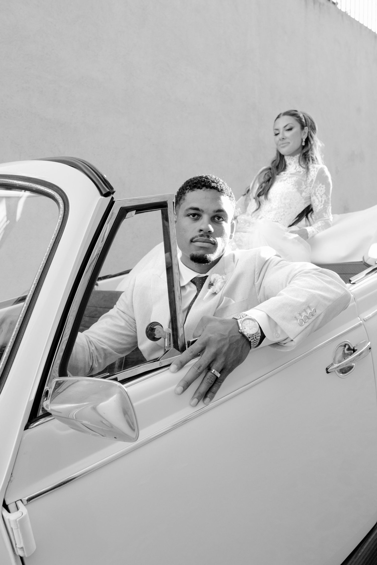 Black and white photograph of bride and groom posing elegantly with a classic vintage car.