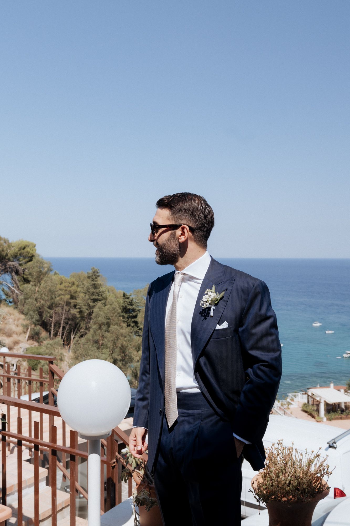 A groom in a navy suit looks out over an ocean view from a scenic coastal wedding venue with white decor.