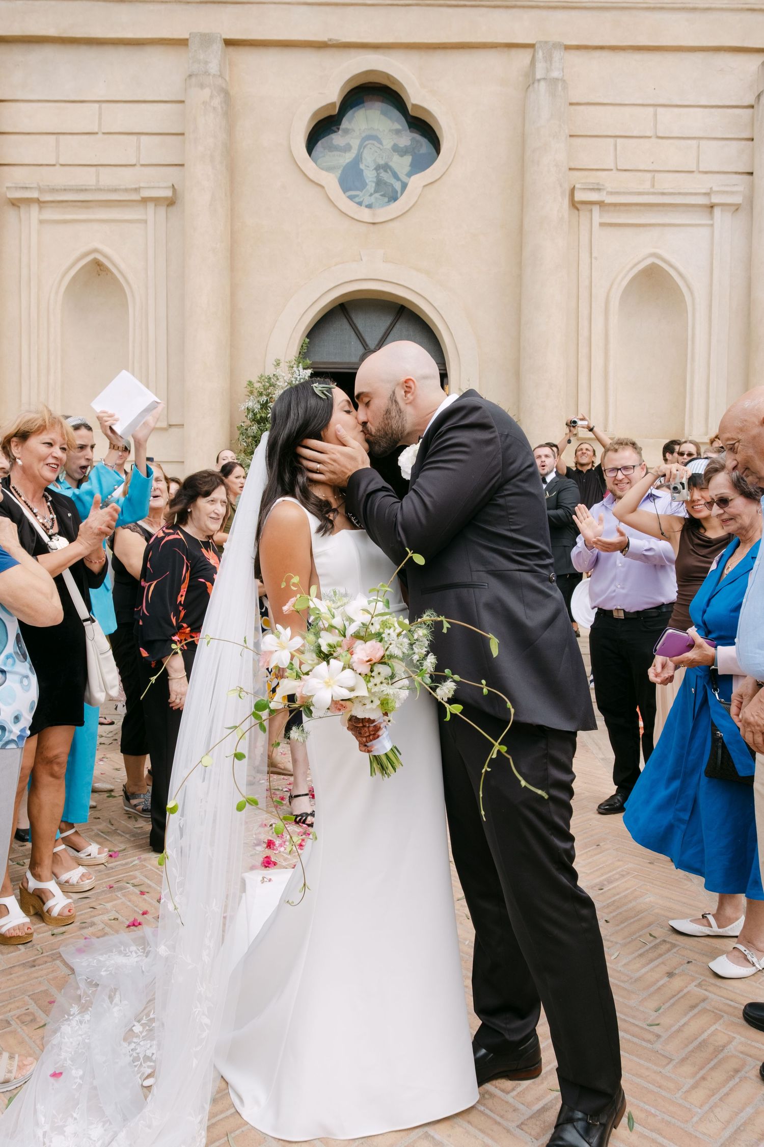 Wedding guests celebrating and throwing flower petals outside a grand stone church.