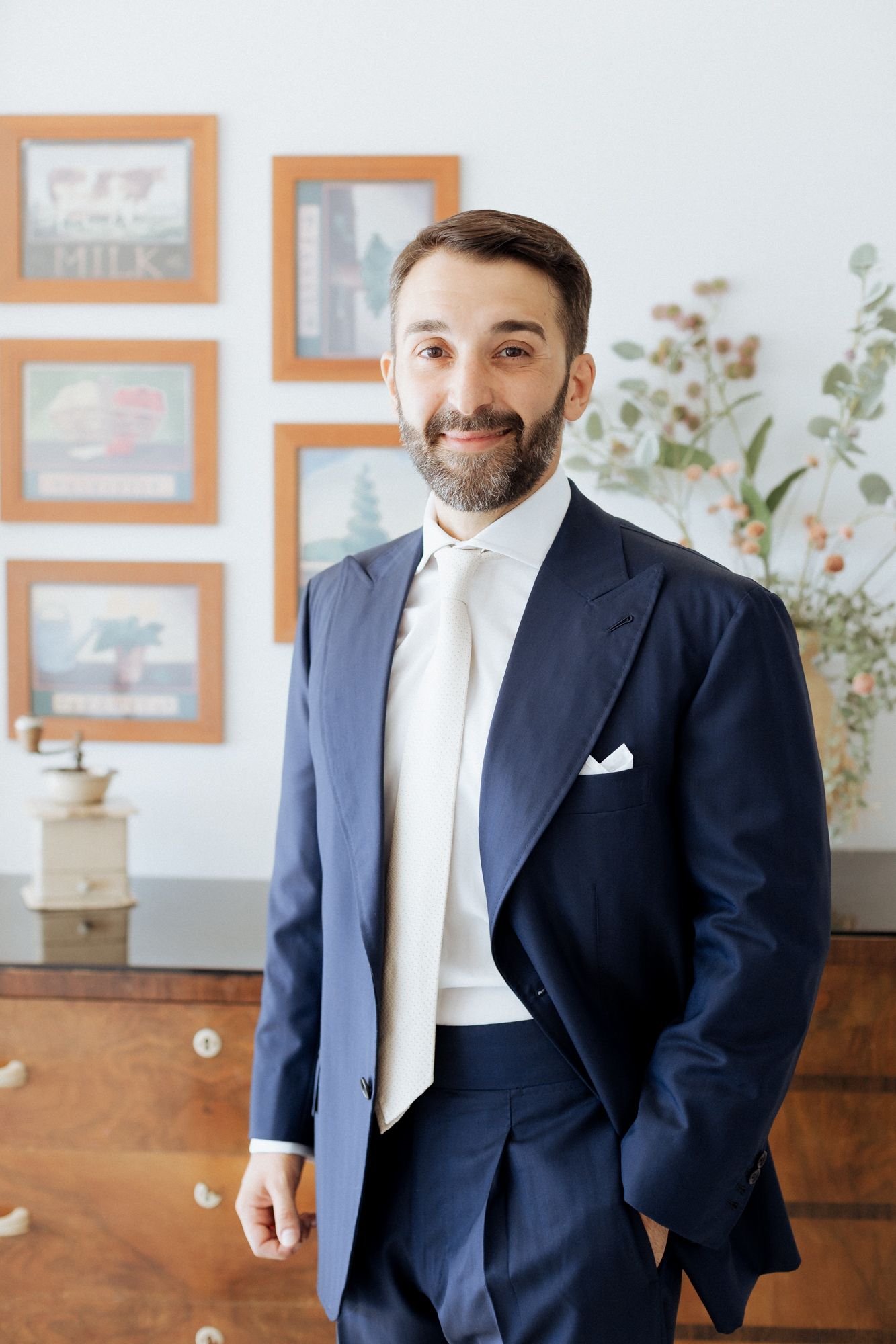 A groom in a navy suit and white tie stands smiling in front of framed artwork and a wooden dresser.