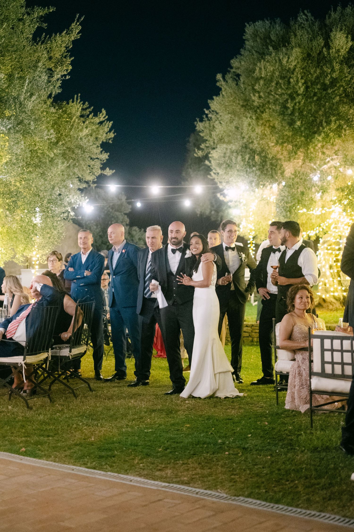 Outdoor nighttime wedding reception with guests gathered under string lights and trees.