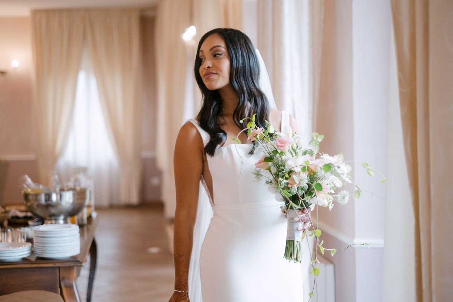 A bride in an elegant white wedding dress holds a delicate bouquet of white and blush flowers against a light backdrop.