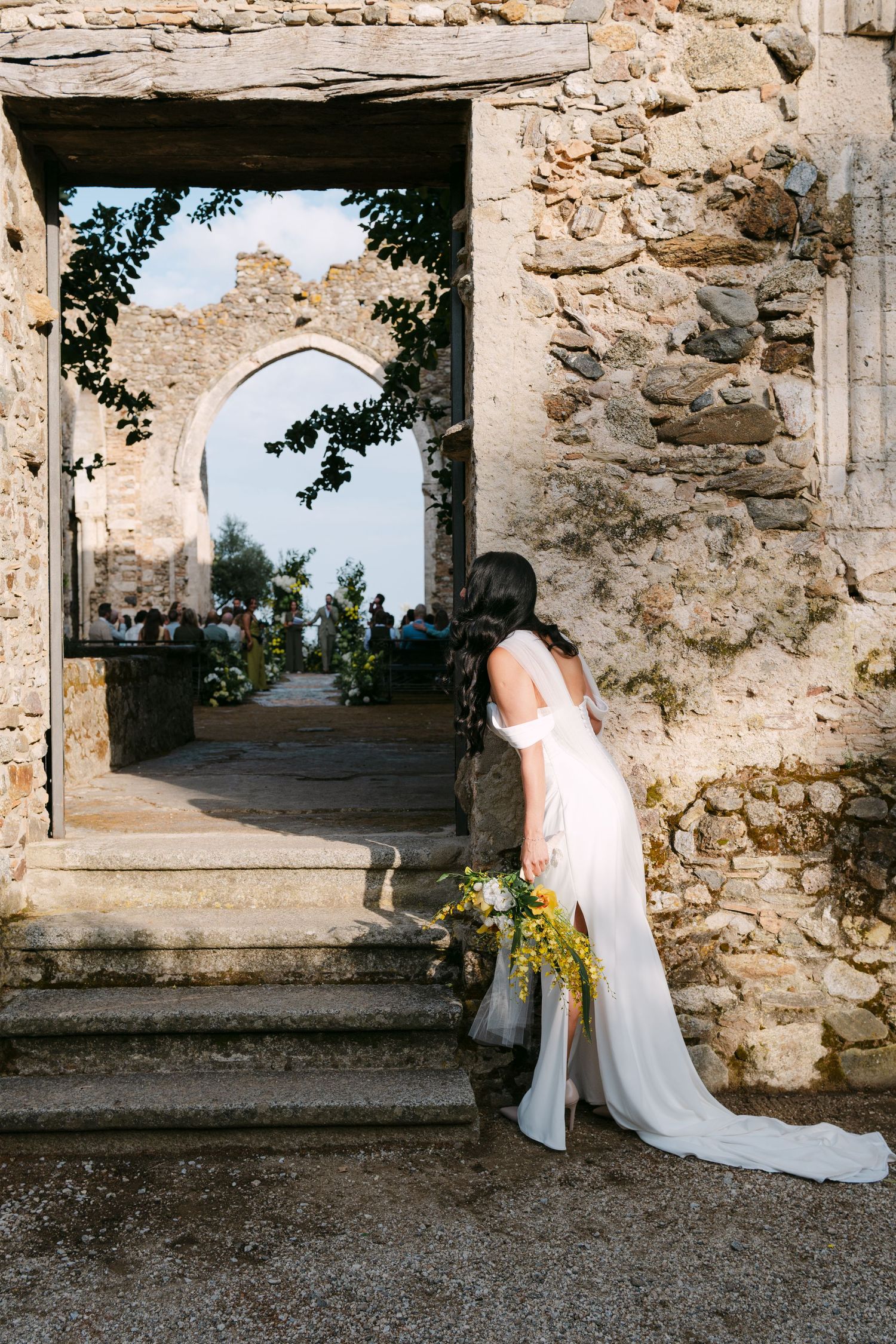 A bride with a bouquet ascends stone steps through an ancient archway.