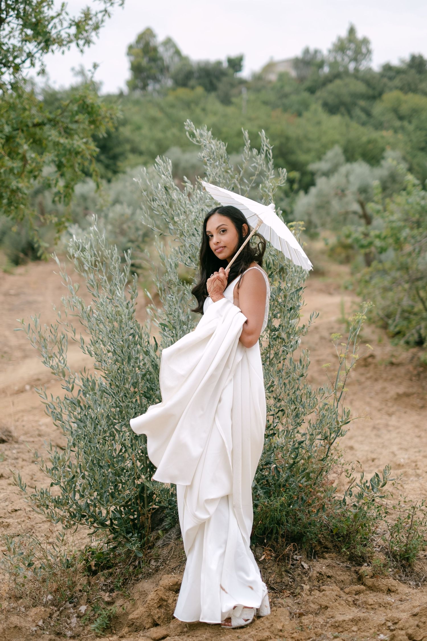 Flowing white wedding dress captured in motion among olive trees in natural setting.