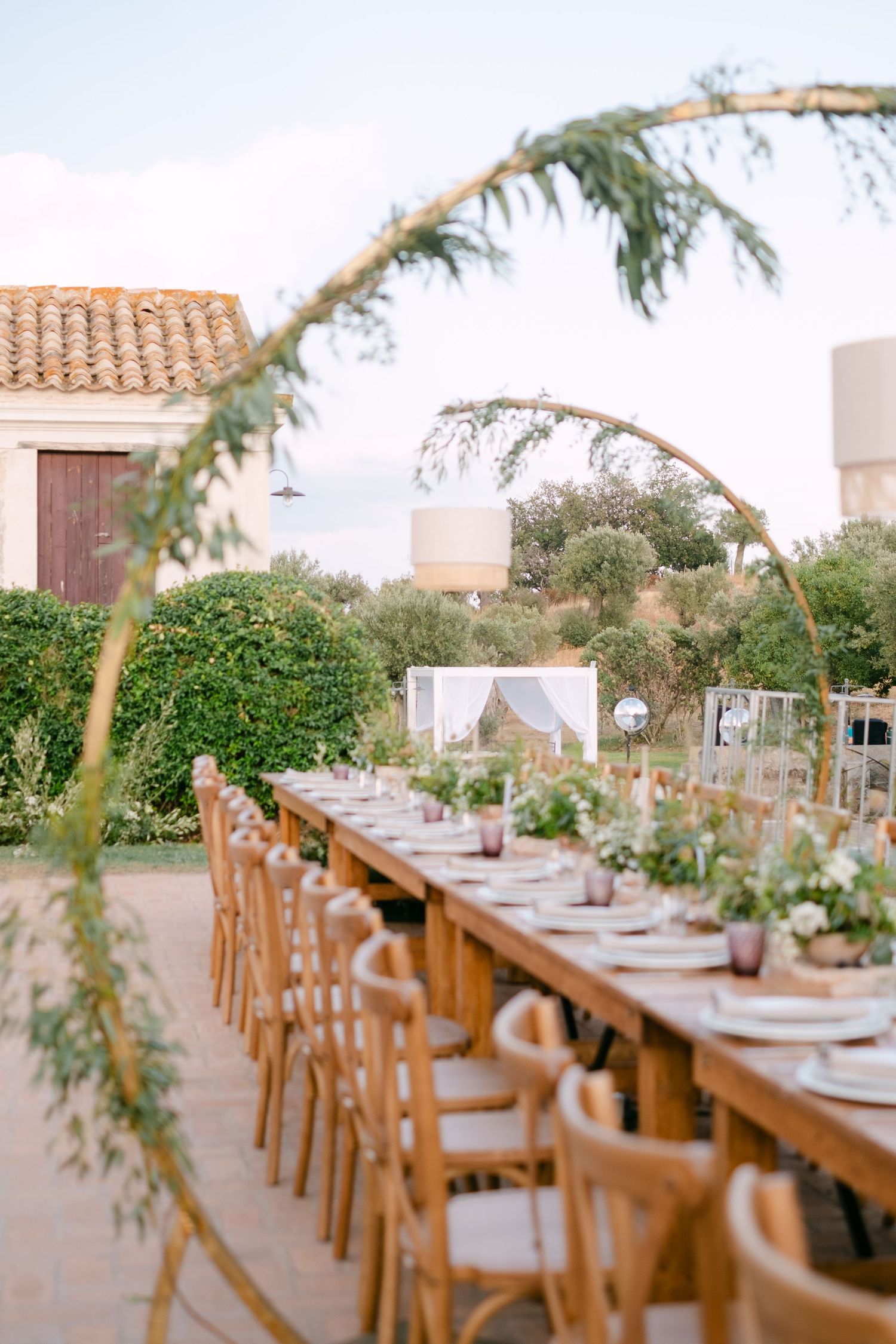 Long rustic dining table set for a wedding reception with wooden chairs and greenery decorations.
