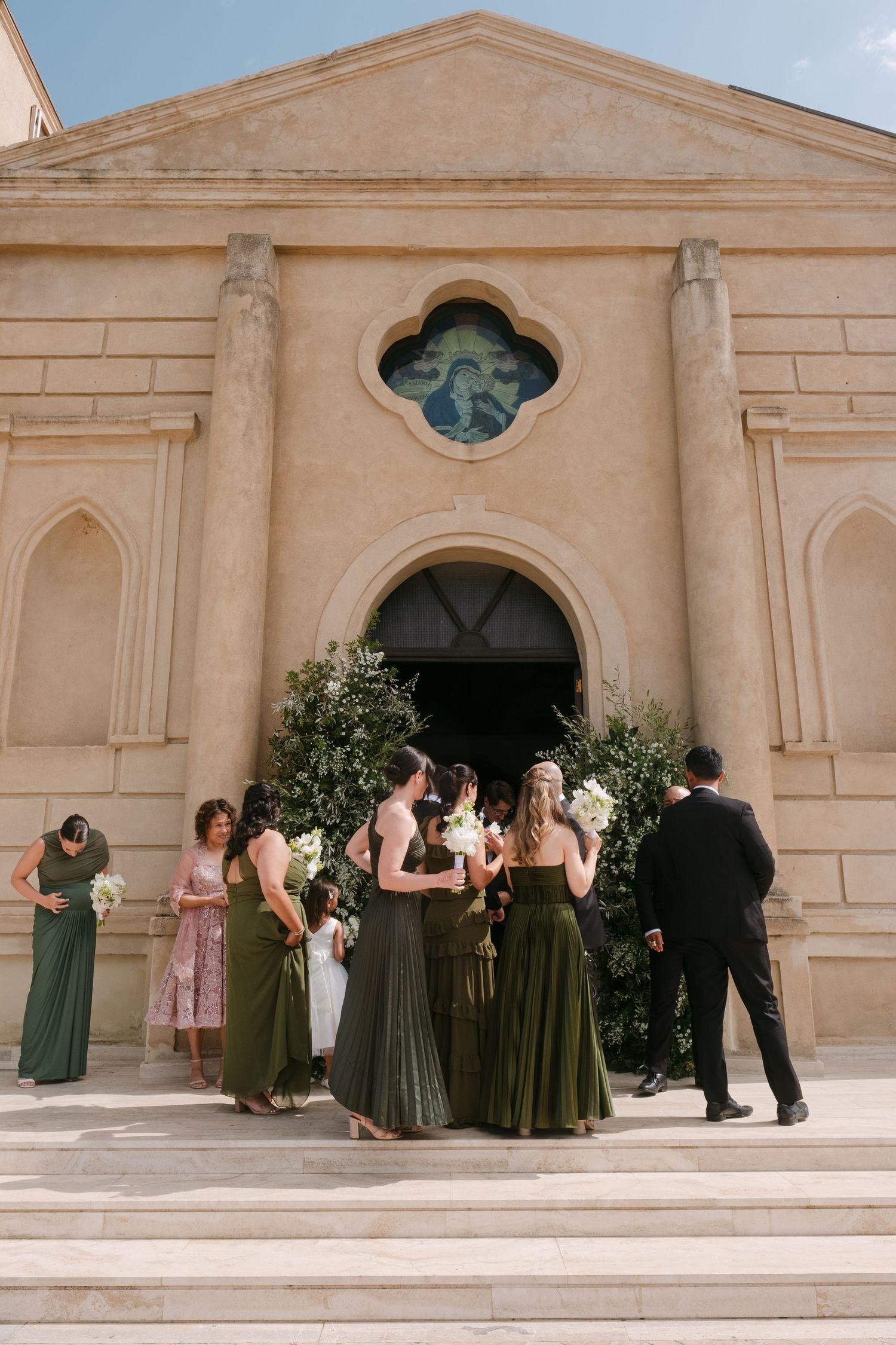 Wedding party gathered on stone church steps with elegant olive green bridesmaid dresses.
