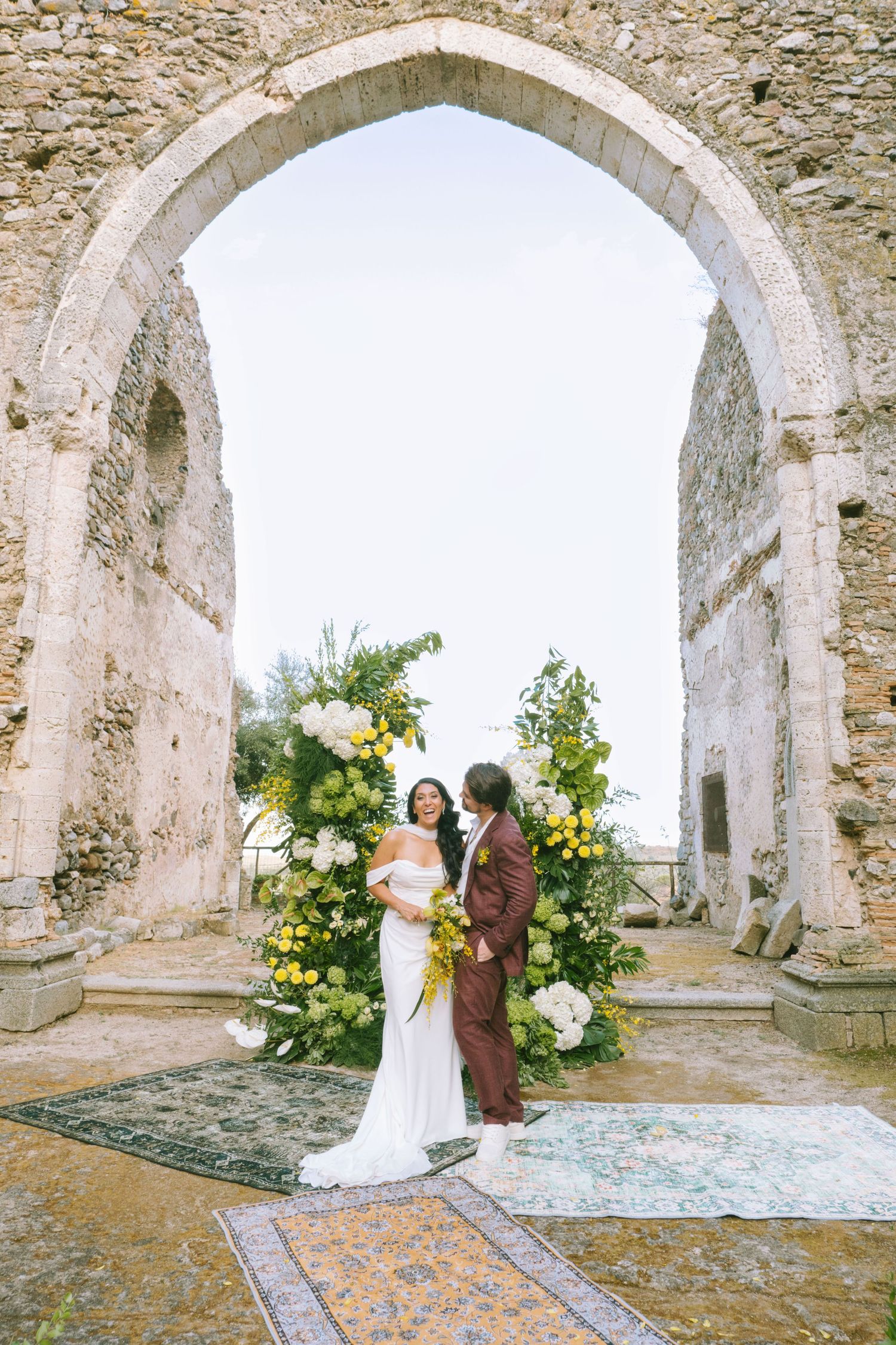 Wedding couple poses under stone archways with elegant floral arrangements in a rustic European setting.