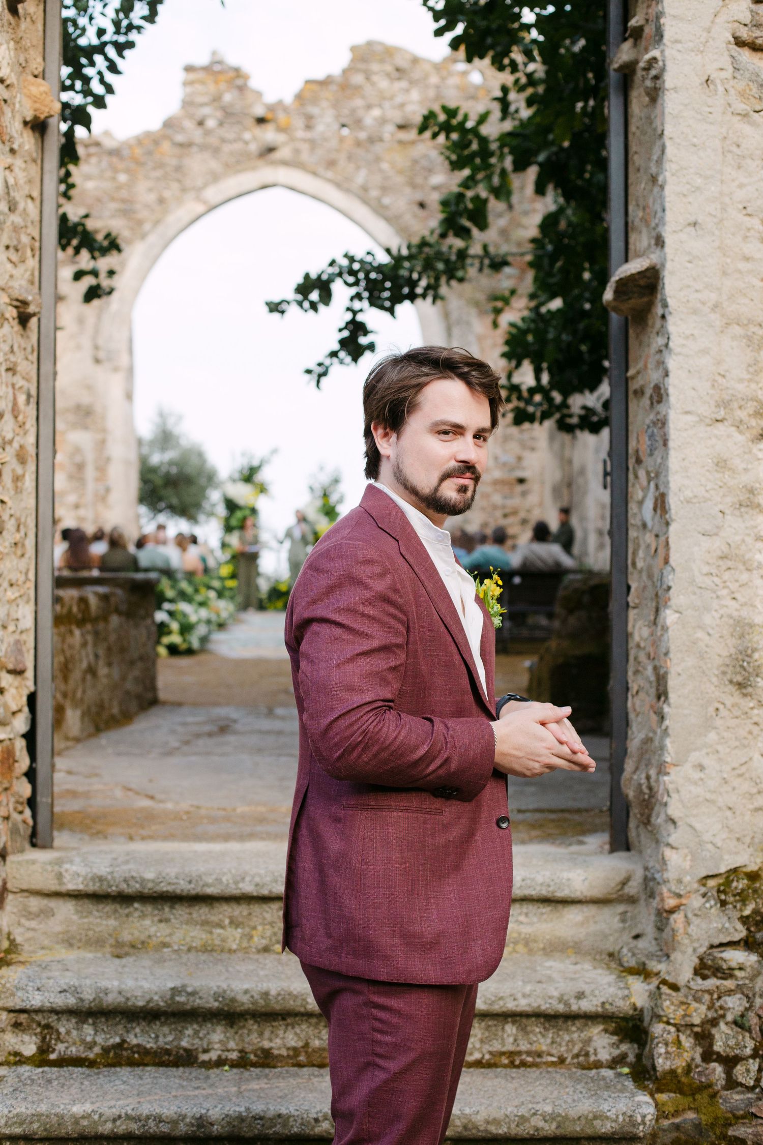 A person in a burgundy suit stands thoughtfully in an ancient stone corridor with crumbling walls and arches.