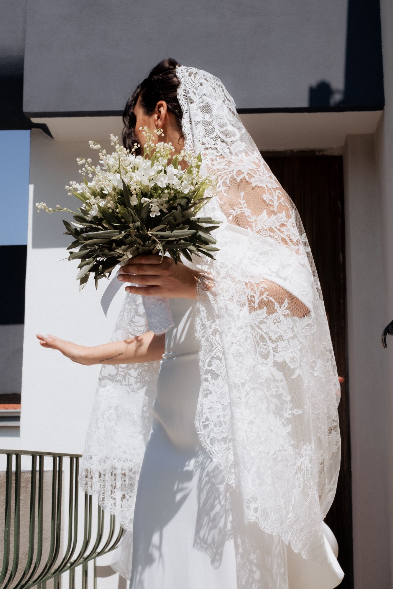 A bride in an elegant lace wedding dress holds a white floral bouquet on a balcony.