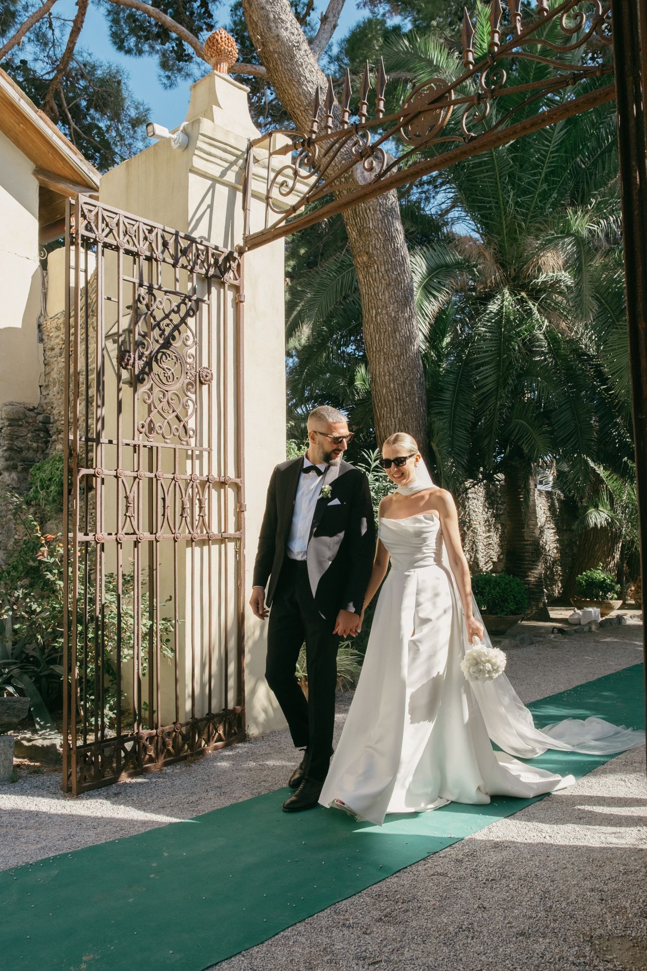 A couple in wedding attire walk through an elegant white gate entrance with trees and greenery in the background.