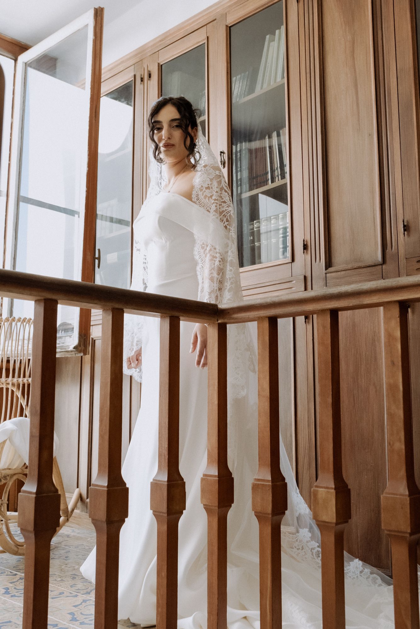 Bride in elegant white lace wedding dress standing behind wooden railing.