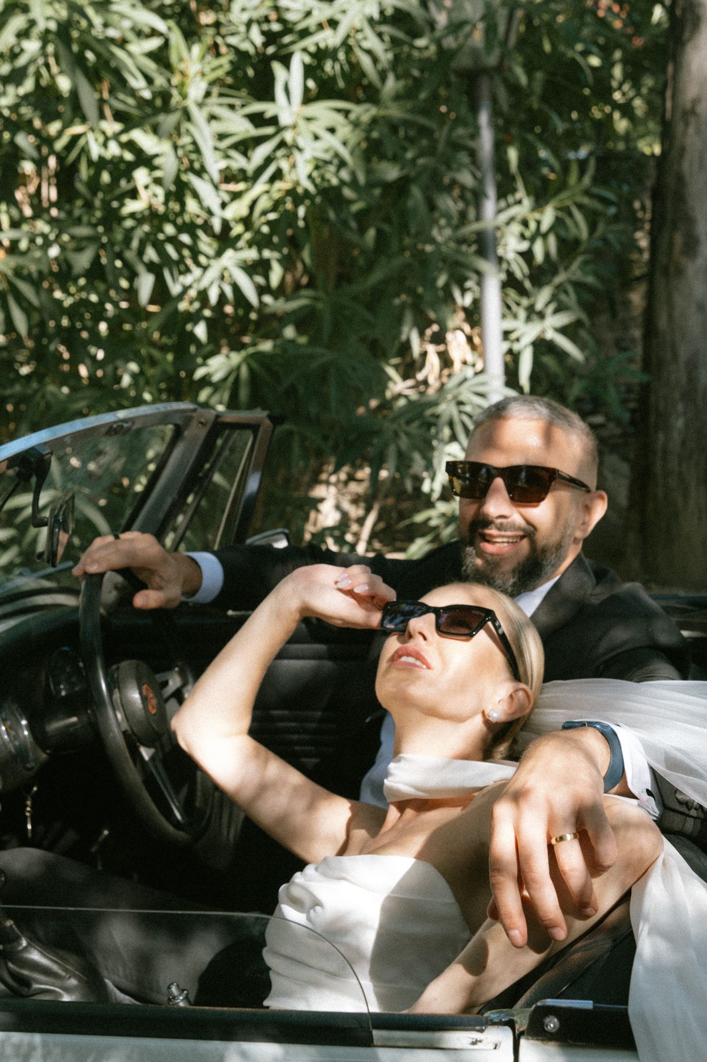 Person in sunglasses drinks from a bottle while relaxing in a car with trees visible in the background.