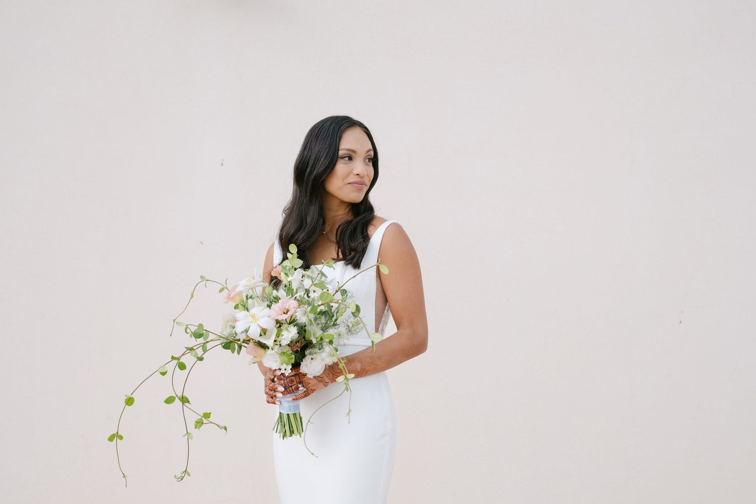 Bride in elegant white sleeveless wedding dress holds cascading bouquet of white flowers and greenery against neutral backdrop.