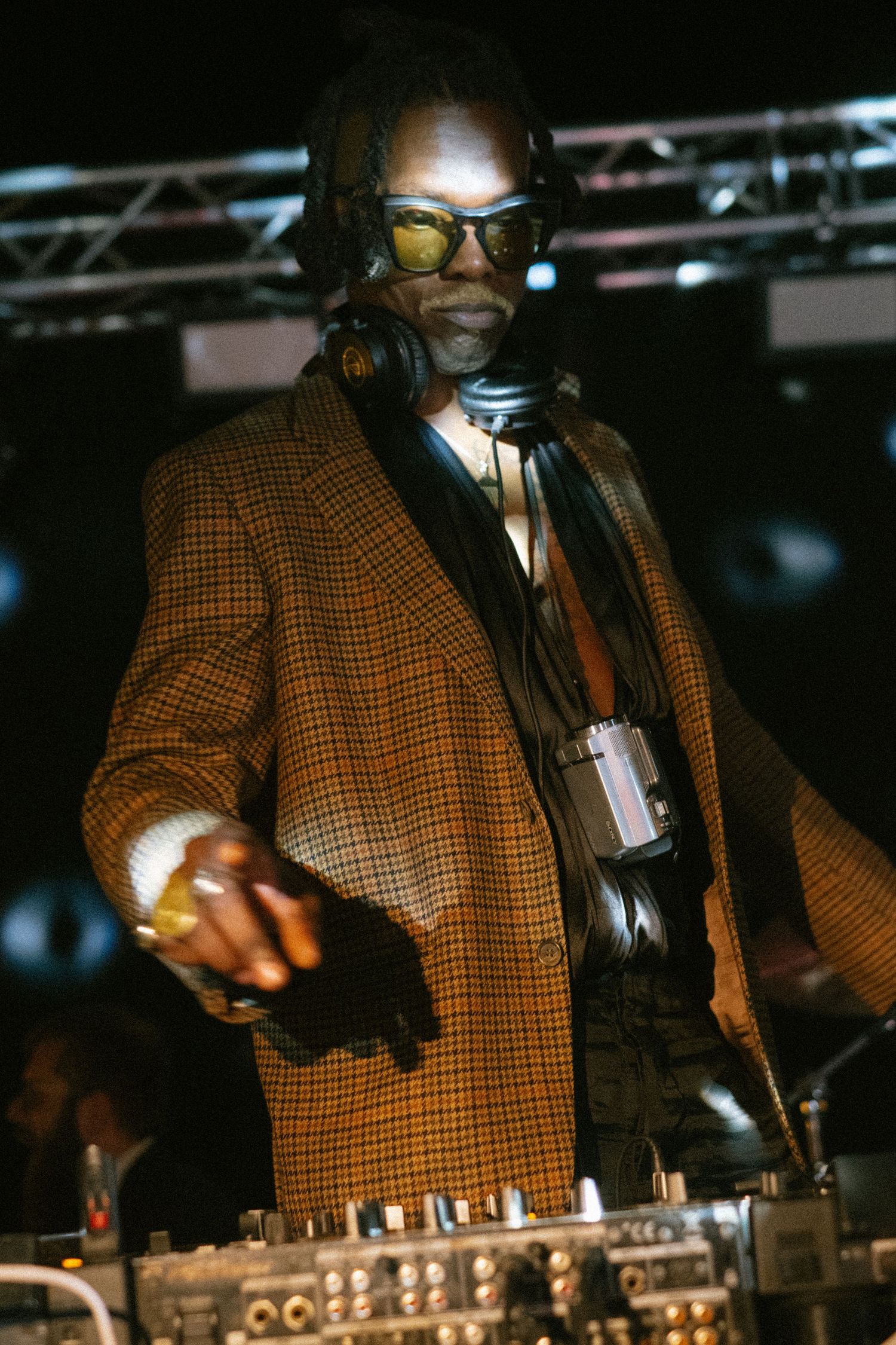 DJ performing at an event wearing a brown checkered jacket while mixing on professional equipment.