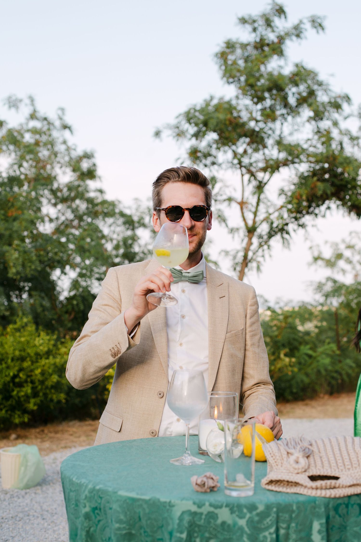 A person in a beige suit and sunglasses enjoys drinks at an outdoor table with a green tablecloth.